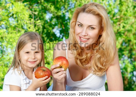 Showing off. Couple of mother and daughter sitting on cover during picnic and holding apples.