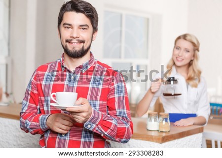 Happy customer. Young man with beard holding cup of coffee on background of waitress.