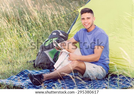 Green tourism.  Portrait of a young handsome tourist wearing blue t-short and beige shorts, sitting on a plaid holding a map looking at the camera smiling, green tent on the background