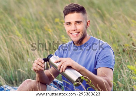 Green tourism.  Portrait of a young handsome tourist wearing blue t-short and beige shorts, sitting on the plaid drinking tea from thermos