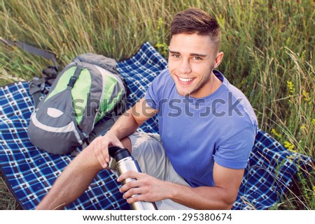 Green tourism.  Top view of a young handsome tourist wearing blue t-short and beige shorts, sitting on the plaid drinking tea from thermos , backpack near