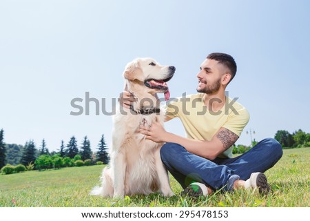 Portrait of a handsome man wearing yellow t-short and jeans with tattoo on his arm sitting on the grass, looking smiling on his lovely golden retriever in the park
