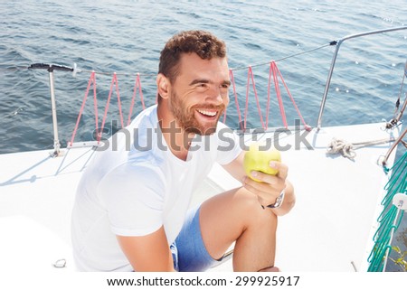Live life fully. Upbeat smiling man sitting on the deck and holding apple while reveling in sailing.