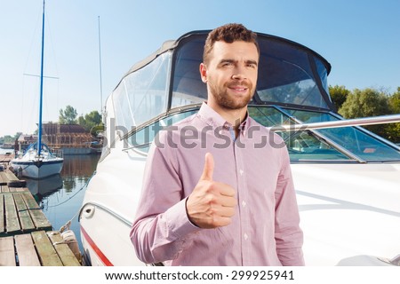 Feeling over the moon. Nice bearded man thumbing up and evincing positivity while standing near his yacht