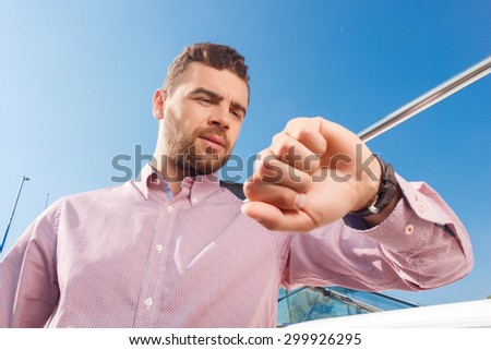 Low angle of attractive man raising his hand and looking at wrist watch while standing near yacht.