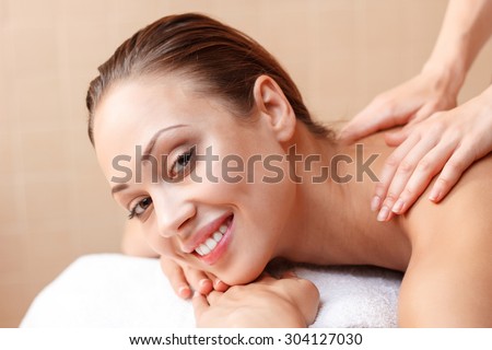 Feeling contented. Pleasant smiling woman lying on the couch and folding arms while professional massager making her massage.