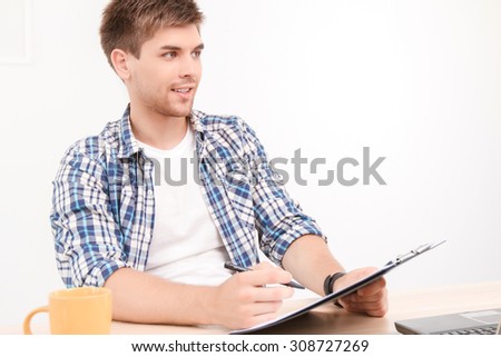 Portrait of a young handsome student sitting on the table holding a folder and a pen looking aside, thinking of new ideas for his project work, in a white room