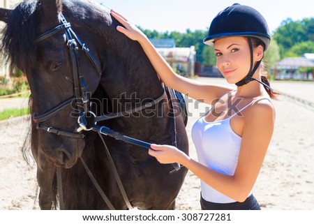 Portrait of a beautiful horsewoman wearing an equestrian helmet and a white top standing near a big brown horse, looking at the camera