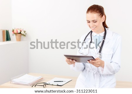 In pace of life. Cheerful smiling beautiful woman doctor holding laptop and standing near table while being busy at work