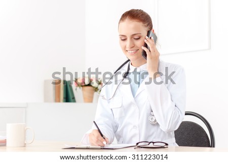 Nice conversation. Contented overjoyed woman doctor writing and talking on mobile phone while sitting at the table