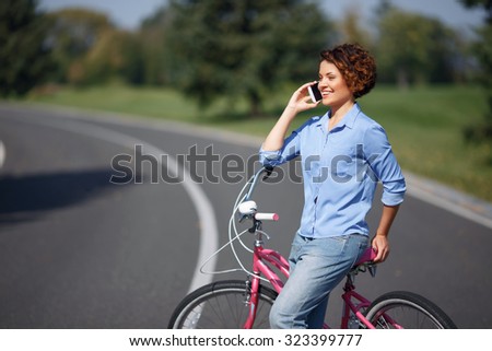 Be active. Nice blissful upbeat young girl holding her bicycle and having brisk conversation on mobile phone while standing on the road