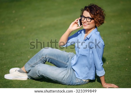 Active girl. Cheerful young attractive girl having conversation on mobile phone and evincing joy while resting on the grass