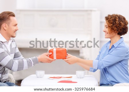 Wish you all the best. Overjoyed handsome man giving present to his girl friend sitting at the table and having dinner.