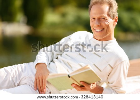 Full  of joy. Agreeable handsome  overjoyed man holding book and reading it while  resting on the blanket