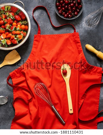 Red kitchen apron on black chalkboard background. Top view. Strawberries and cherries on a dark background. Apron and berries. Space for text. The summer berries. Preparation of baking from berries.
