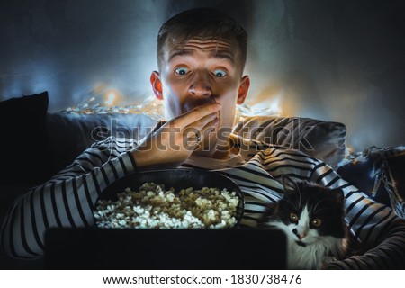 young man with black cat watching a movie eating popcorn on TV at home. Movie night. Relax,rest watching a horror film or video on screen. Background lighting. Fun Scared excited people on the couch. Stock fotó © 