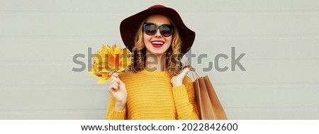Autumn portrait of happy smiling young woman with shopping bags and yellow maple leaves wearing a sweater and hat on gray background
