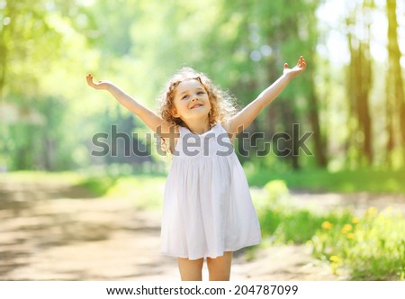 Charming little girl enjoying summer sunny day, raises his hands top, expression emotions, happy child walking in the park