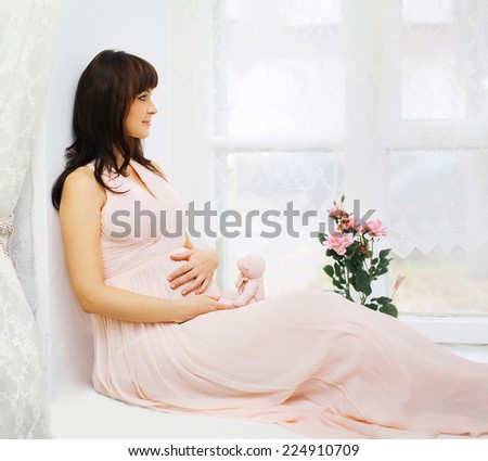 Pregnancy, motherhood and happy future mother concept - beautiful pregnant woman in dress home, dreams of a future child looking in window