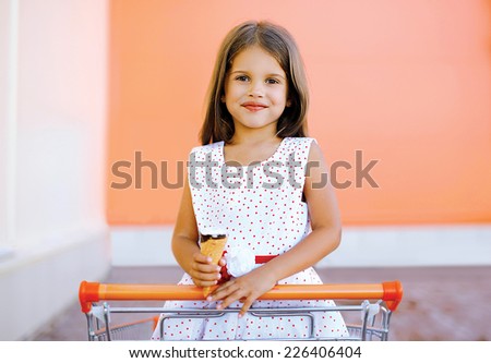 Portrait happy little girl in shopping cart with tasty ice cream having fun in the city