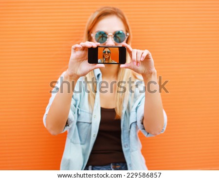 Summer, fun, technology and people concept - cool girl makes self-portrait on the smartphone outdoors against colorful wall