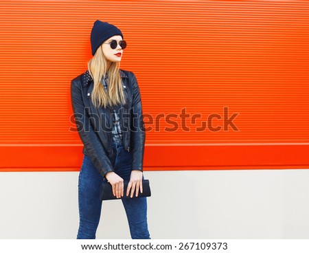 Fashion portrait of stylish blonde girl in rock black style, wearing a sunglasses and leather jacket with clutch standing outdoors against the red urban wall in the city