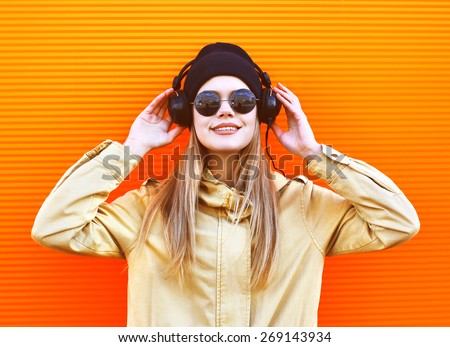 Portrait stylish woman wearing a black hat and headphones listens to music enjoys freedom, cool hipster girl in the city against a colorful orange wall, street fashion concept, urban style