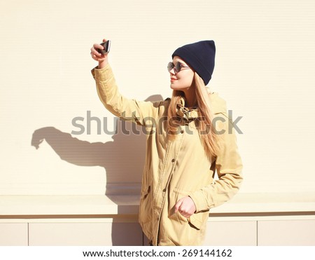 Fashion pretty blonde makes selfie-portrait on the smartphone, carefree hipster girl wearing a black sunglasses and hat, enjoying and having fun outside in sunny day against the urban wall