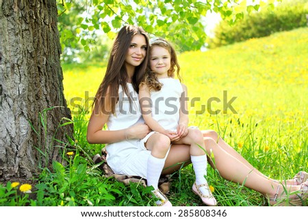 Mother and daughter together on the grass near tree in summer day, mom with child outdoors
