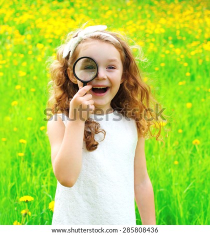 Portrait of positive little girl looking through a magnifying glass on summer floral field