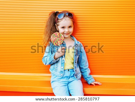 Fashion kid - stylish little girl child wearing a jeans clothes and sunglasses with sweet caramel having fun in city and enjoys summer against the colorful orange wall