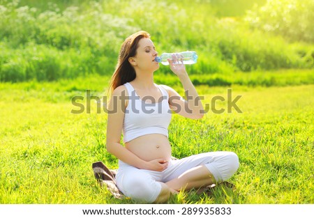 Pregnancy, sport and health lifestyle - young pregnant woman drinking water from a bottle sitting on the grass in sunny summer day