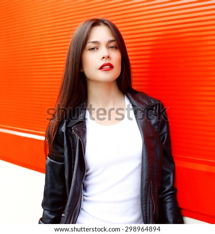 Fashion portrait of sexy beautiful brunette woman with red lipstick wearing a rock black leather jacket outdoors in the city against the colorful wall