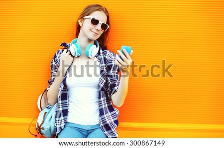 Pretty girl in sunglasses and headphones using smartphone against the colorful orange wall