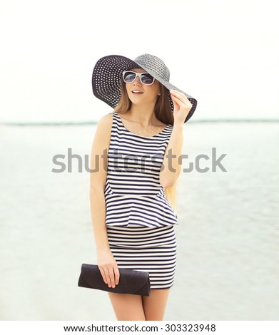 Fashion young woman wearing a striped dress and summer black straw hat with handbag clutch over sea