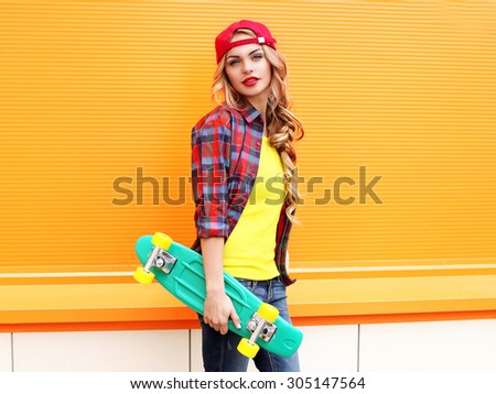 Fashion portrait of pretty young woman in red checkered hipster shirt, cap with skateboard against the colorful orange background