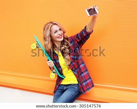 Pretty smiling blonde young woman in colorful clothes with skateboard having fun makes self-portrait on the smartphone against the orange background