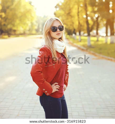 Fashion blonde woman wearing a sunglasses and red leather jacket with scarf in autumn park