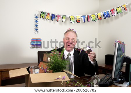 Senior man in a business suit sitting behind a desk with retirement banner, box full of personal effects holding a cupcake