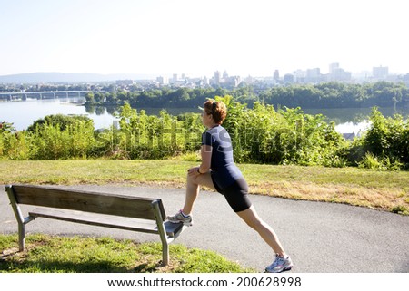 Female jogger stretching on a bench before running along a path overlooking a river view city skyline on a hazy, hot summer day.