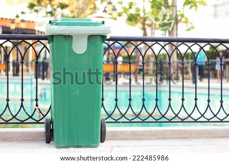 Green plastic trash through use, located along the fence in the park.