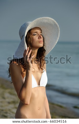 young woman in a white bikini on a bach wearing a white sun hat with her eyes closed