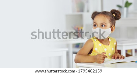 A dark-skinned girl in a medical mask for respiratory protection in a yellow dress listens attentively to the teacher. white background. Banner format