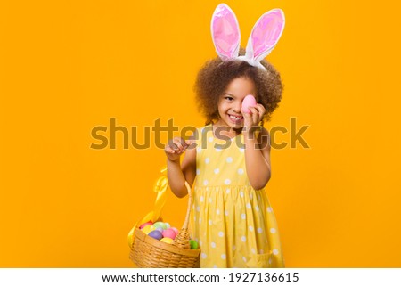 A cheerful girl with rabbit ears on her head with a basket of colored eggs in her hands on a yelow background. Funny crazy happy baby. Easter to the child.