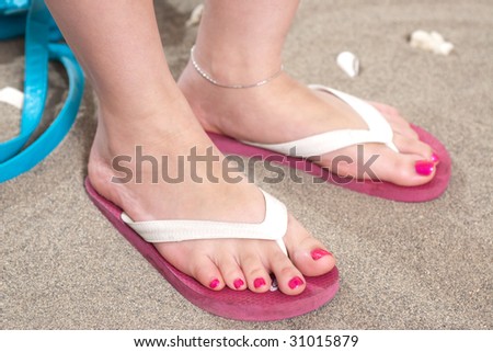 A woman\'s feet  in pink flip flops on the sand with a pedicure and pink nail polish.