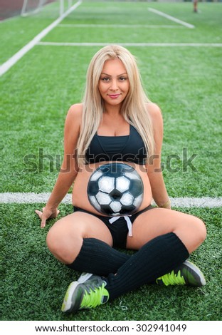 attractive pregnant woman with soccer ball on the belly sitting on the football field