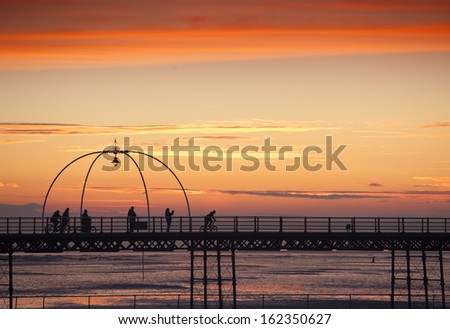 Silhouetted figures at sunset on Southport Pier. One figure taking a photograph another walking and some riding bicycles.