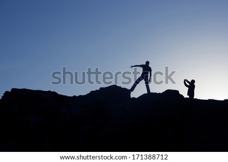 Silhouettes of two men standing on rocks one pointing and one holding camera taking photograph isolated against blue sky.