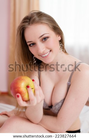 portrait of sexy blonde girl having fun happy smiling holding big red apple sitting on white bed in lingerie and looking at camera on light window copy space background