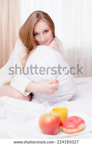 breakfast in bed: portrait of beautiful sexy blonde young lady lying on white bed and happy smiling looking at camera on light copy space background